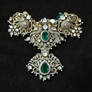 The image shows a close-up of a jewelry piece, likely a pendant or a necklace. The jewelry is adorned with a large, pear-shaped gemstone, possibly a diamond or a moissanite, surrounded by a halo of smaller gemstones. The setting appears to be made of silver or white gold, with intricate floral designs and filigree work. The background is a dark, velvety material.