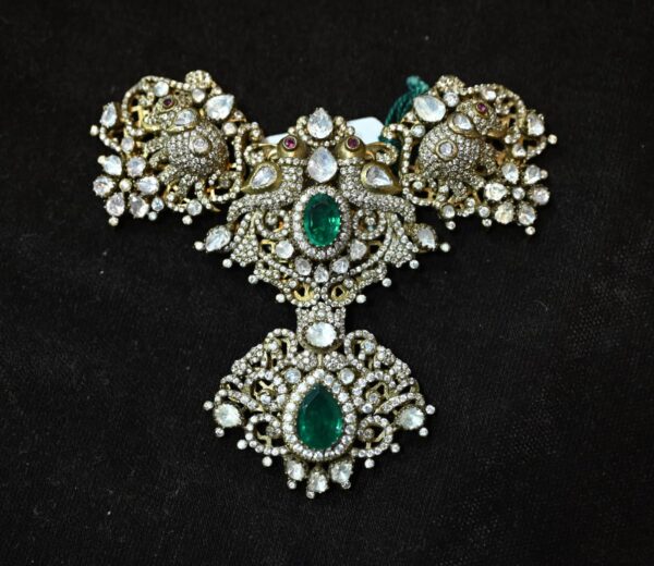 The image shows a close-up of a jewelry piece, likely a pendant or a necklace. The jewelry is adorned with a large, pear-shaped gemstone, possibly a diamond or a moissanite, surrounded by a halo of smaller gemstones. The setting appears to be made of silver or white gold, with intricate floral designs and filigree work. The background is a dark, velvety material.
