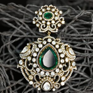 The image shows a close-up of a delicate earring. It appears to be a dangle earring with a pear-shaped green gemstone as the centerpiece. The gemstone is surrounded by a cluster of smaller, colorless stones, creating a halo effect. The earring is further embellished with intricate filigree work, adding a touch of vintage charm. The background is a dark, textured surface, which contrasts beautifully with the sparkle of the earring.