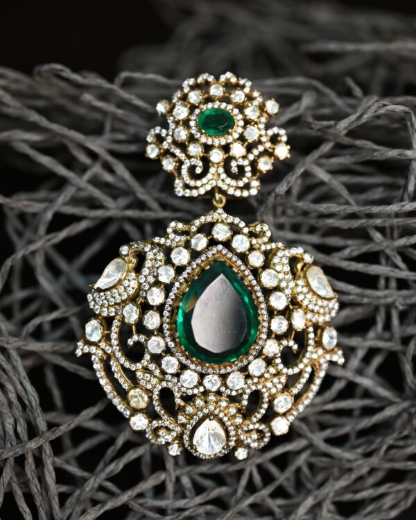 The image shows a close-up of a delicate earring. It appears to be a dangle earring with a pear-shaped green gemstone as the centerpiece. The gemstone is surrounded by a cluster of smaller, colorless stones, creating a halo effect. The earring is further embellished with intricate filigree work, adding a touch of vintage charm. The background is a dark, textured surface, which contrasts beautifully with the sparkle of the earring.