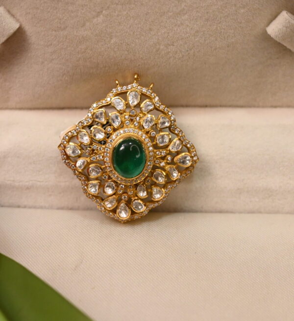 The image shows a beautiful pendant featuring a vibrant green moissanite gemstone. The stone is set in a delicate gold-tone mount and surrounded by a halo of smaller moissanite stones. The overall design of the pendant has a classic floral motif. The pendant is displayed in a jewelry box, with a red tulip placed beside it. The contrast of the green stone against the gold and the red flower creates a visually appealing composition.