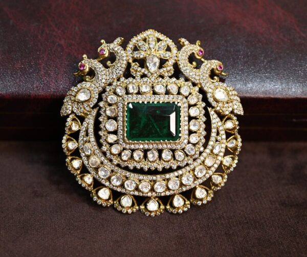 The image shows a close-up of a beautiful jewelry pendant. The pendant is circular in shape and is adorned with a large, square-cut emerald moissanite stone in the center. The emerald moissanite is surrounded by a halo of smaller, round-cut moissanite stones. The pendant also features intricate filigree work and two delicate peacock motifs on either side, adding a touch of elegance and whimsy to the design. The pendant appears to be set in gold.