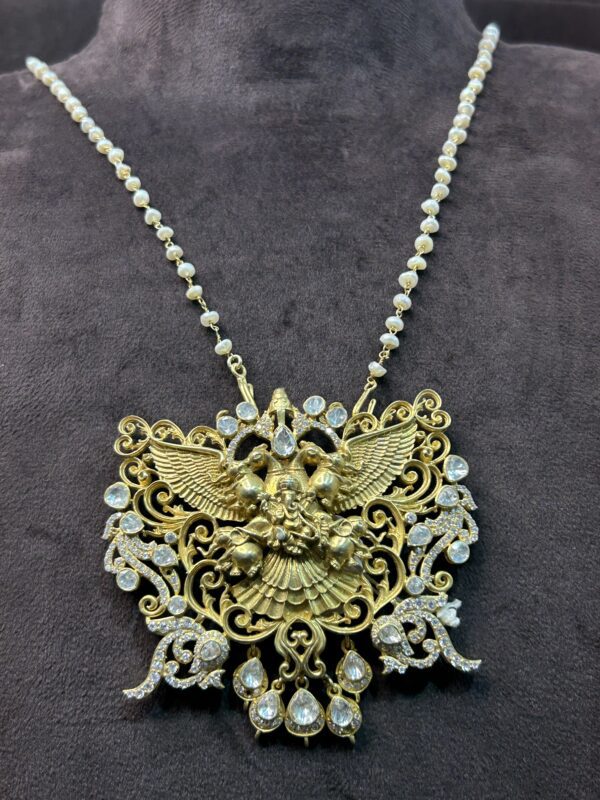The image you shared is a close-up of a gold pendant depicting a Hindu deity. The pendant is intricately crafted with delicate details and features a combination of gold and pearls. The deity is shown with multiple arms, each holding a different symbol or weapon. The pendant is suspended from a gold chain.