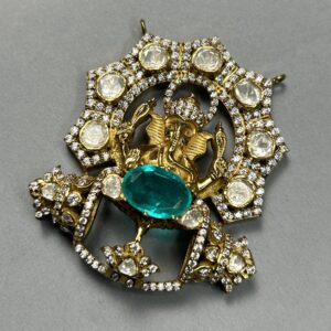 The image shows a close-up of a jewelry piece, likely a pendant or a brooch. It appears to be made of gold and is heavily embellished with sparkling stones. The central design features a raised depiction of Lord Ganesha, the Hindu god of wisdom and prosperity. Ganesha is shown seated on a throne and is adorned with intricate details. The background of the pendant is filled with smaller stones, creating a shimmering effect.