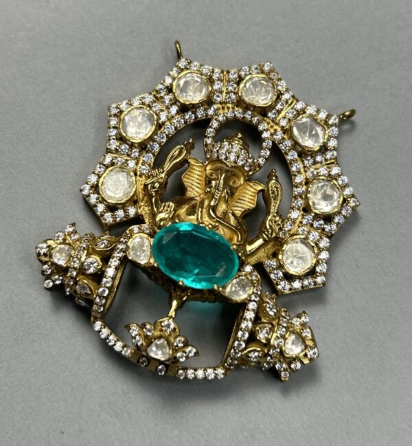 The image shows a close-up of a jewelry piece, likely a pendant or a brooch. It appears to be made of gold and is heavily embellished with sparkling stones. The central design features a raised depiction of Lord Ganesha, the Hindu god of wisdom and prosperity. Ganesha is shown seated on a throne and is adorned with intricate details. The background of the pendant is filled with smaller stones, creating a shimmering effect.