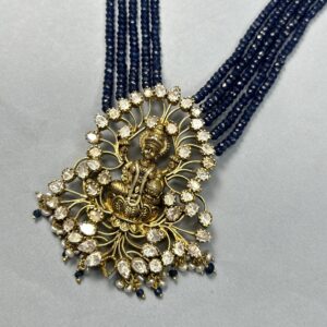 The image shows a stunning necklace with a striking centerpiece. The necklace itself is composed of multiple strands of small, deep blue beads, creating a rich and luxurious look. The centerpiece is a large, ornate pendant featuring a gold-toned deity figure, likely Lord Krishna or Vishnu, seated in a meditative pose.