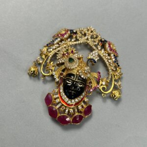 The image shows a beautiful silver temple pendant. It appears to be a depiction of Shrinathji, a form of Krishna worshipped in Nathdwara, Rajasthan. The pendant is adorned with intricate details and colorful gemstones, adding to its visual appeal and spiritual significance.