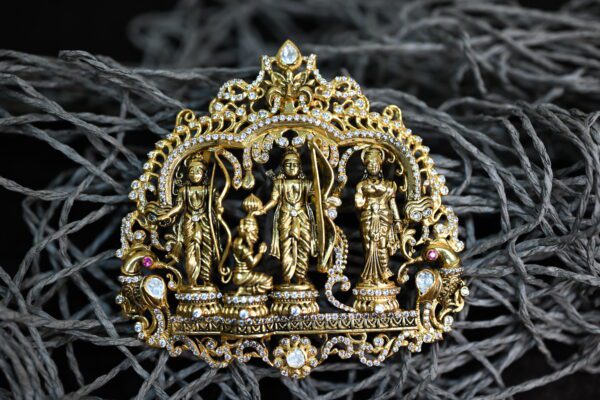 The image showcases a close-up of a silver temple pendant. The pendant features an intricate design depicting the Hindu deity Lord Ram, seated with his wife Sita and brother Lakshman. Hanuman, the devoted monkey god, is standing reverently before them. The pendant is adorned with sparkling white stones and pink gemstones, enhancing its elegance.