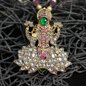 The image shows a beautiful temple pendant featuring Goddess Lakshmi, the Hindu goddess of wealth, prosperity, and good fortune. The pendant is crafted with intricate details and features a combination of precious and semi-precious s tones. The background of the image is a textured surface, possibly a piece of fabric or a natural material, which provides a contrast and highlights the intricate details of the pendant.