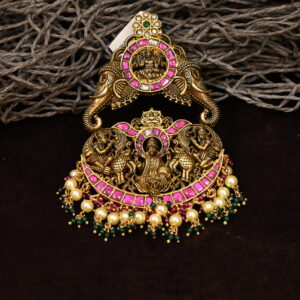 The image showcases a stunning temple-style pendant. It appears to be crafted from a gold-tone metal and is intricately designed with a central motif depicting a Hindu deity. The pendant is embellished with vibrant pink and green gemstones, adding a touch of color and sparkle.