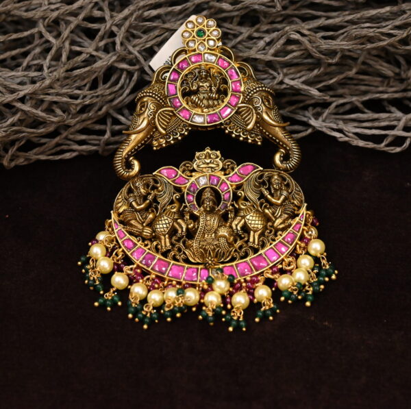 The image showcases a stunning temple-style pendant. It appears to be crafted from a gold-tone metal and is intricately designed with a central motif depicting a Hindu deity. The pendant is embellished with vibrant pink and green gemstones, adding a touch of color and sparkle.