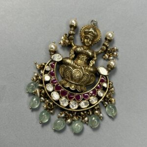The image shows a close-up of a delicate gold pendant. The pendant features a central figure, likely a Hindu deity, seated gracefully on a lotus flower. The deity is adorned with intricate details and appears to be wearing elaborate jewelry.