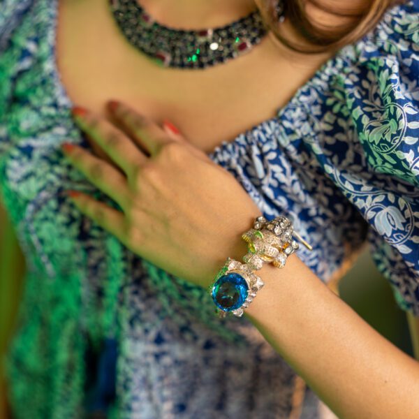 The image shows a close-up of a woman's wrist wearing an elaborate diamond bangle. The bangle features a large, round, blue gemstone surrounded by a halo of smaller diamonds. Adjacent to this is another decorative element, possibly gold-toned and also encrusted with numerous diamonds, perhaps in a stylized floral or animal shape. The two main sections are connected by a band that also appears to be set with diamonds.