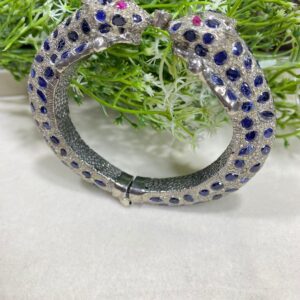 The image shows an ornate bracelet designed in the shape of two leopards facing each other. The bracelet is predominantly silver-toned and is encrusted with blue and clear stones, forming a spotted pattern similar to that of a leopard's fur. The eyes of the leopards are made with pink stones, adding a vivid contrast to the rest of the design. Blue Sapphire Pave Diamonds Bangle