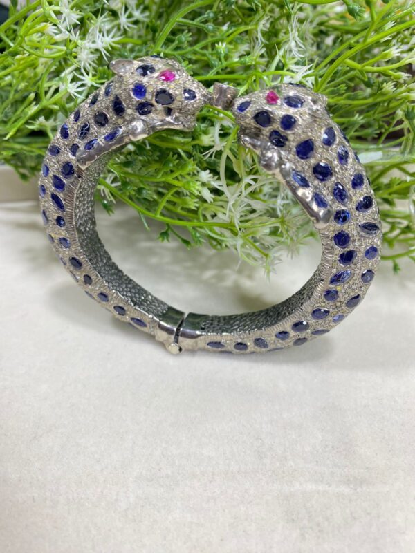 The image shows an ornate bracelet designed in the shape of two leopards facing each other. The bracelet is predominantly silver-toned and is encrusted with blue and clear stones, forming a spotted pattern similar to that of a leopard's fur. The eyes of the leopards are made with pink stones, adding a vivid contrast to the rest of the design. Blue Sapphire Pave Diamonds Bangle