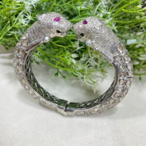 The image showcases a luxurious, intricately designed bracelet with two panther heads facing each other at the ends. The panther heads are encrusted with dazzling white stones, giving them a sparkling appearance, and each has a striking pink gemstone for an eye. The bracelet's band features a detailed pattern, with a mix of smooth and textured elements that add depth and elegance. The entire piece is silver-toned, likely made of white gold or platinum. The background includes green foliage and delicate white flowers, adding a natural, contrasting backdrop that enhances the brilliance and opulence of the jewelry. Premium Uncut Diamond Bangle