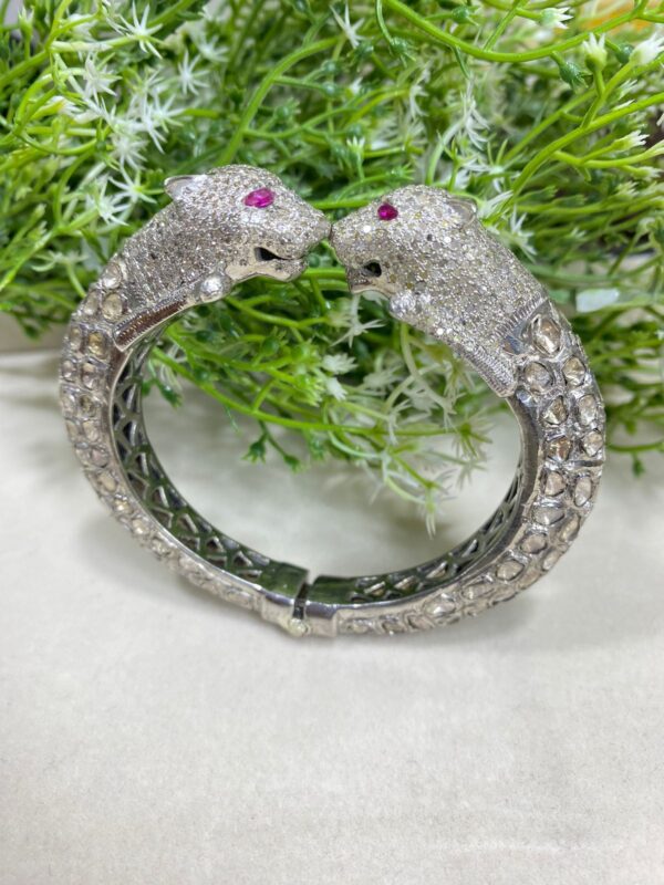The image showcases a luxurious, intricately designed bracelet with two panther heads facing each other at the ends. The panther heads are encrusted with dazzling white stones, giving them a sparkling appearance, and each has a striking pink gemstone for an eye. The bracelet's band features a detailed pattern, with a mix of smooth and textured elements that add depth and elegance. The entire piece is silver-toned, likely made of white gold or platinum. The background includes green foliage and delicate white flowers, adding a natural, contrasting backdrop that enhances the brilliance and opulence of the jewelry. Premium Uncut Diamond Bangle