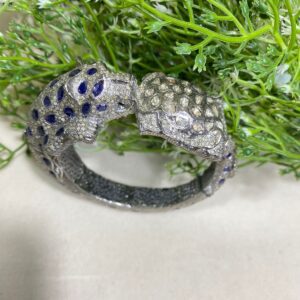 The image shows an ornate bracelet designed in the shape of leopard facing each other. The bracelet is predominantly silver-toned and is encrusted with blue and clear stones, forming a spotted pattern similar to that of a leopard's fur. The eyes of the leopards are made with pink stones, adding a vivid contrast to the rest of the design. Blue Sapphire Diamonds Bangle