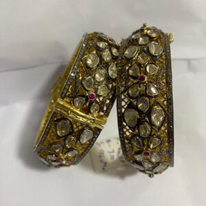 This image showcases a pair of ornate, likely Indian, bangles. They appear to be crafted from 925 sterling silver with gold accents and feature a rich embellishment of natural diamonds. The diamonds are predominantly in the Polki cut, retaining their organic shapes, and possibly some smaller Rosecut diamonds. Adding color are small, round pave-set rubies (red) and blue sapphires. The metalwork exhibits an intricate, possibly openwork design with areas of darker, oxidized silver contrasting with the lighter tones. These bangles exude a vintage or antique charm, indicative of traditional Indian jewelry with potential historical influences. Unique Pave Diamond Bangle