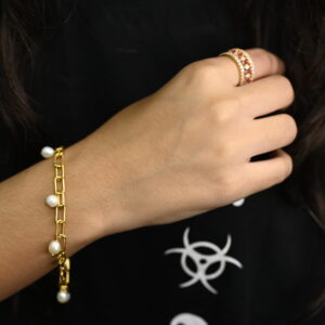 A hand models a delicate gold-plated 925 silver bracelet adorned with small pearls and a matching ring set with rubies and smaller stones. The jewelry provides a subtle yet elegant accent against a dark background featuring a white symbol.