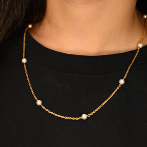 The image shows a person wearing a delicate necklace. The necklace appears to be made of a thin, solid gold chain. At evenly spaced intervals along the chain are small, white pearls. The design is minimalist and elegant. The necklace rests against the backdrop of a plain black garment. The focus is sharply on the jewelry, highlighting its simplicity and the contrast between the gold and the pearls. The wearer has dark hair visible in the upper left corner of the frame. The overall impression is one of understated sophistication.