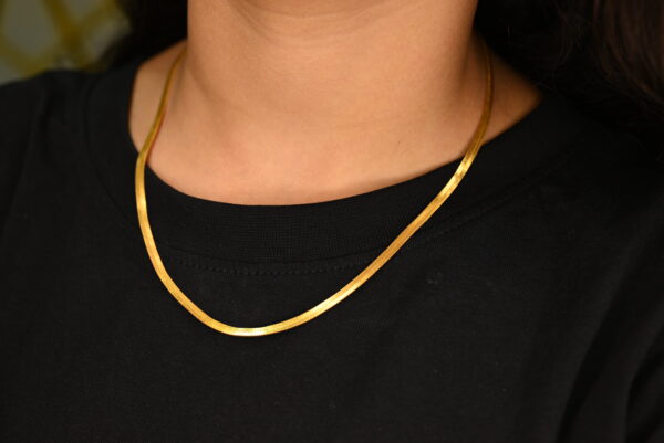 This image shows a close-up of a person wearing a simple yet elegant gold-plated chain necklace. The necklace has a flat, smooth, and shiny design, laying neatly against a black t-shirt. The warm gold tone contrasts beautifully with the black fabric, highlighting the necklace's sleek and polished appearance. The focus is on the neck and necklace, with no visible facial features, emphasizing the jewelry as the main subject.