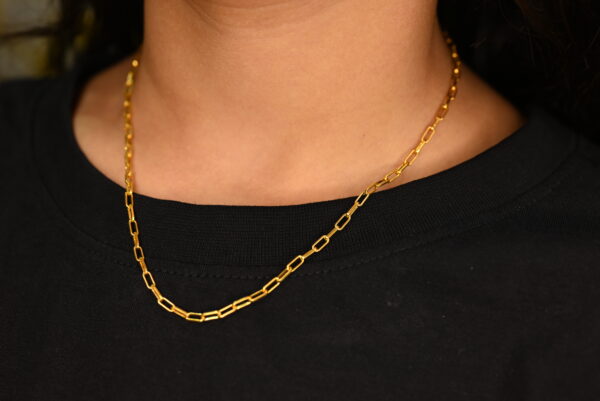A close-up shot shows a delicate, minimalist necklace made of solid gold links resting on a person's neck wearing a plain black t-shirt. The necklace is the clear focal point, highlighting its simple yet elegant design.