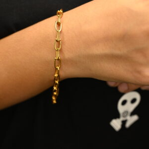A close-up shot showcases a minimalist gold-plated chain bracelet on a wrist. The bracelet features small, elongated oval links and rests gently against the skin.