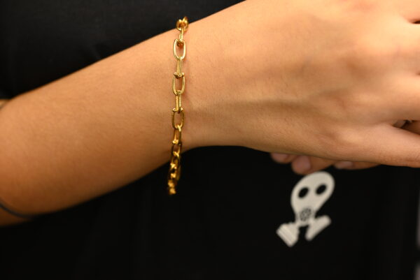 A close-up shot showcases a minimalist gold-plated chain bracelet on a wrist. The bracelet features small, elongated oval links and rests gently against the skin.