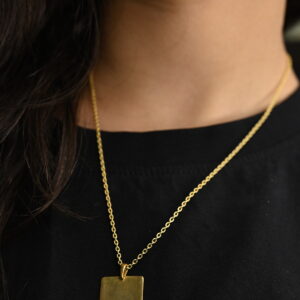 A close-up shows a person wearing a delicate gold necklace with a small, rectangular solid gold pendant. The pendant is engraved with the words "THE WORLD IS YOURS". The necklace rests against a black garment. Minimal 925 Silver Pendant