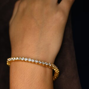 The image shows a close-up of a wrist wearing a delicate gold tennis bracelet. The bracelet is crafted from a warm-toned yellow gold and features numerous small, clear, and sparkling stones, likely diamonds or cubic zirconia, set closely in a continuous line. These stones catch the light, creating a subtle yet noticeable shimmer. The bracelet wraps elegantly around the wrist, suggesting a comfortable and secure fit. The skin tone of the arm is light to medium, with fine hairs visible. The background is blurred and dark, drawing full attention to the jewelry. The overall impression is one of understated luxury and classic style.