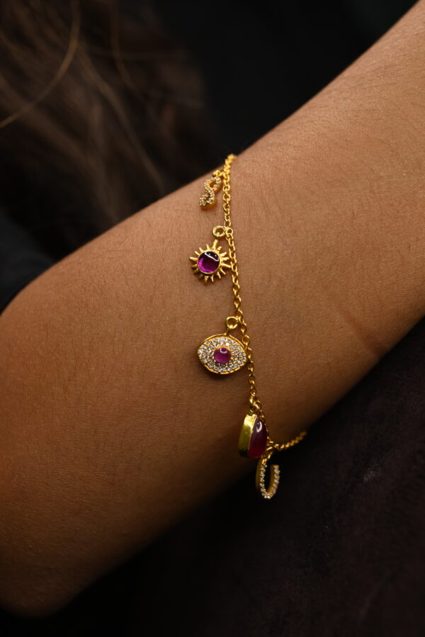 This image shows a delicate gold-plated chain bracelet worn on a person's wrist. The bracelet is made of 925 sterling silver and features several small hanging charms. Each charm is beautifully detailed — some are studded with tiny clear stones surrounding deep pink or ruby-colored center stones, while others are simple polished gems. The chain is fine and minimalistic, enhancing the elegance of the piece. The background is dark, making the gold and jewel tones stand out vividly against the skin. The overall impression is one of refined luxury and subtle sophistication.