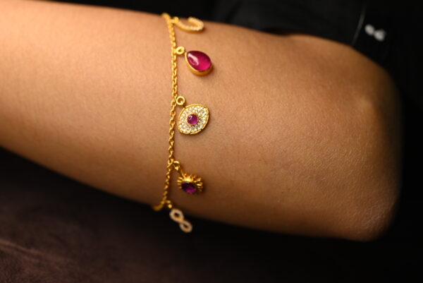 This image shows a delicate gold-plated chain bracelet worn on a person's wrist. The bracelet is made of 925 sterling silver and features several small hanging charms. Each charm is beautifully detailed — some are studded with tiny clear stones surrounding deep pink or ruby-colored center stones, while others are simple polished gems. The chain is fine and minimalistic, enhancing the elegance of the piece. The background is dark, making the gold and jewel tones stand out vividly against the skin. The overall impression is one of refined luxury and subtle sophistication.