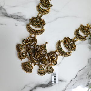 The image shows a traditional South Indian temple-style necklace laid out on a white marble surface with green decorative plants on the right. The necklace is made of antique gold-finished metal and features intricate craftsmanship. At the center is a large pendant of Goddess Lakshmi, seated on a lotus and flanked by two elephants, symbolizing prosperity and divine blessings. Surrounding the pendant are fan-shaped and crescent motifs adorned with green and white stones, adding vibrant detail. The necklace has a rich, ornate appearance typical of temple jewelry, ideal for festive or ceremonial wear. A barcode tag is also visible at the bottom, indicating it may be new or for sale.
