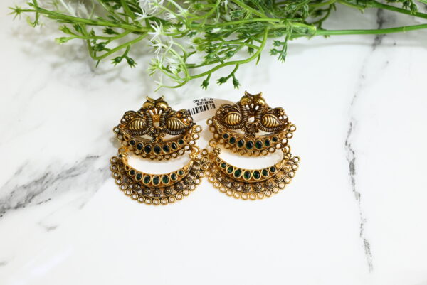 The image showcases a pair of traditional Indian temple-style earrings placed on a white marble surface with green foliage in the background. The earrings are made of 925 silver with an antique gold finish. Each earring features intricate detailing, including two peacocks at the top, floral motifs in the center, and a crescent-shaped lower half adorned with green gemstones and ornate circular patterns along the edge. A price tag reading is partially visible behind the earrings. The overall aesthetic is rich, cultural, and elegant, suitable for ethnic or festive wear.