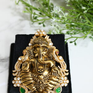 The image showcases an intricately designed temple-style pendant featuring Lord Ganesha, crafted in a gold-finished 925 silver. The deity is depicted in a traditional seated posture with finely detailed ornaments and expressions. The pendant is adorned with sparkling white stones and highlighted with green gem-like accents near the base. It is placed on a black velvet box, enhancing its rich and regal appearance. In the background, soft greenery adds a serene and spiritual touch to the composition, making the pendant stand out as a divine and artistic piece of jewelry.