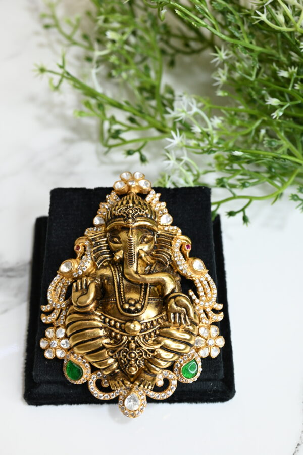 The image showcases an intricately designed temple-style pendant featuring Lord Ganesha, crafted in a gold-finished 925 silver. The deity is depicted in a traditional seated posture with finely detailed ornaments and expressions. The pendant is adorned with sparkling white stones and highlighted with green gem-like accents near the base. It is placed on a black velvet box, enhancing its rich and regal appearance. In the background, soft greenery adds a serene and spiritual touch to the composition, making the pendant stand out as a divine and artistic piece of jewelry.