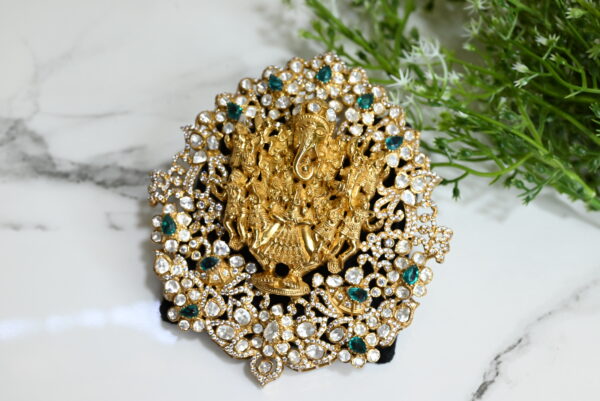 The image shows an intricately designed temple-style pendant placed on a white marble surface with green foliage in the background. The pendant is made from 925 silver and plated with gold. At the center, it features a highly detailed depiction of Lord Ganesha, surrounded by celestial figures in traditional attire, symbolizing divine presence and protection. The outer frame of the pendant is studded with a combination of white and green stones, arranged in floral and teardrop patterns, enhancing its opulence and traditional charm. The piece reflects exquisite craftsmanship, typical of South Indian temple jewelry.