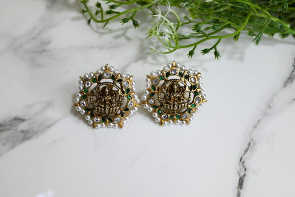 The image showcases a pair of intricately designed traditional temple earrings placed on a white marble surface with a green leafy plant in the background. The earrings are crafted in 925 silver with an antique gold finish and feature a central depiction of Goddess Lakshmi seated in a blessing pose. Surrounding the deity are sparkling white stones and green accents, bordered with a ring of small white pearls. The overall design is circular with a star-like pattern, exuding a regal and divine aesthetic typical of South Indian temple jewelry. The scene is well-lit, emphasizing the rich detailing and craftsmanship of the earrings.