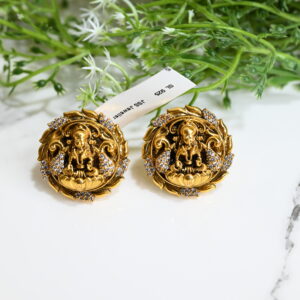 The image showcases a pair of intricately designed temple-style earrings. Made from 925 silver and finished with a rich gold plating, each earring features a central deity figure—likely a Hindu goddess—seated on a lotus. The surrounding design resembles a floral or ornamental frame encrusted with sparkling white stones, likely cubic zirconia, enhancing the overall opulence. The earrings rest on a white marble surface with a lush green plant in the background, and a tag reading "SL 925 JSS Jeweller" is attached, indicating authenticity and craftsmanship. The overall aesthetic is traditional, luxurious, and culturally significant.
