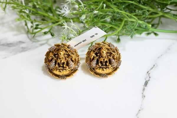 The image showcases a pair of intricately designed temple-style earrings. Made from 925 silver and finished with a rich gold plating, each earring features a central deity figure—likely a Hindu goddess—seated on a lotus. The surrounding design resembles a floral or ornamental frame encrusted with sparkling white stones, likely cubic zirconia, enhancing the overall opulence. The earrings rest on a white marble surface with a lush green plant in the background, and a tag reading "SL 925 JSS Jeweller" is attached, indicating authenticity and craftsmanship. The overall aesthetic is traditional, luxurious, and culturally significant.