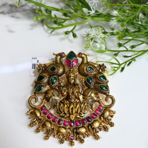The image showcases an intricately designed temple-style pendant placed on a white marble surface with green decorative foliage in the background. The pendant is made of 925 silver with an antique gold finish. At the center, it features a detailed idol of Goddess Lakshmi seated on a lotus, symbolizing wealth and prosperity. Flanking her are two majestic peacocks adorned with green enamel work, and elephants are positioned below, enhancing the regal feel. The pendant is embellished with pink and green gemstones, arranged beautifully to complement the traditional motif. Tiny hanging elephant charms add movement and charm to the overall piece.