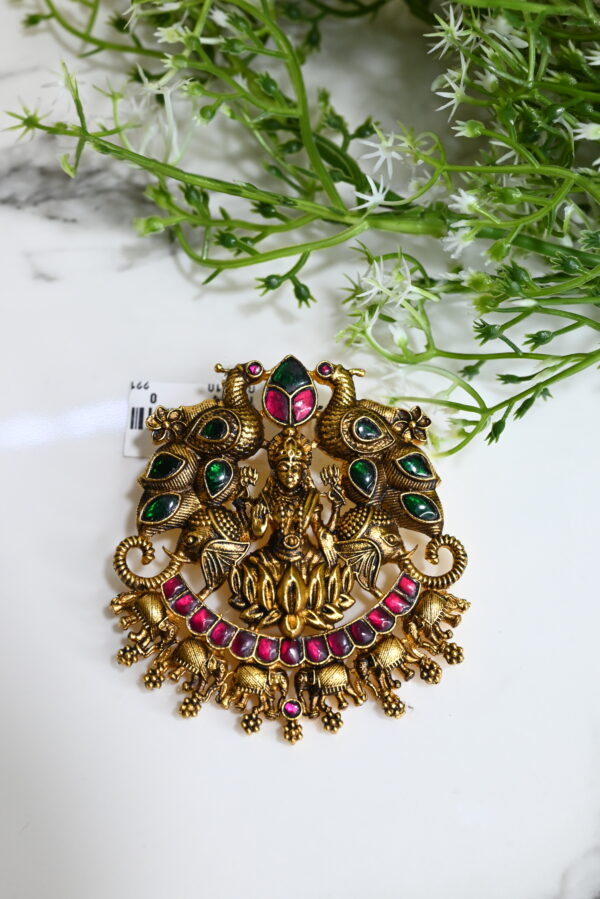 The image showcases an intricately designed temple-style pendant placed on a white marble surface with green decorative foliage in the background. The pendant is made of 925 silver with an antique gold finish. At the center, it features a detailed idol of Goddess Lakshmi seated on a lotus, symbolizing wealth and prosperity. Flanking her are two majestic peacocks adorned with green enamel work, and elephants are positioned below, enhancing the regal feel. The pendant is embellished with pink and green gemstones, arranged beautifully to complement the traditional motif. Tiny hanging elephant charms add movement and charm to the overall piece.