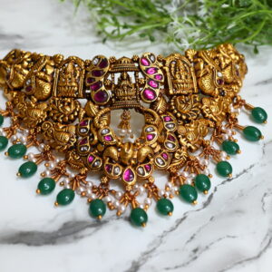The image showcases a stunning traditional temple-style necklace, designed in a rich antique gold finish. The centerpiece features intricate carvings of Hindu deities and motifs such as peacocks and floral patterns, reflecting South Indian craftsmanship. The necklace is adorned with ruby-pink and white kundan stones, adding a vibrant contrast. Dangling from the bottom are clusters of small white pearls paired with teardrop-shaped green beads, enhancing its regal appeal. The necklace is placed on a white marble surface with green foliage in the background, emphasizing its luxurious and cultural essence.