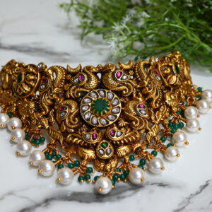 The image showcases an ornate temple-style choker necklace, richly designed in antique gold plating over 925 silver. The centerpiece features a circular green stone surrounded by clear kundan stones. The necklace is adorned with intricate motifs, including peacocks and floral patterns, highlighted with ruby and emerald-colored stones. Hanging below are clusters of white pearls and green beads, adding elegance and movement to the piece. The background is a smooth white marble surface with a sprig of green foliage, enhancing the luxurious and traditional appeal of the jewelry.
