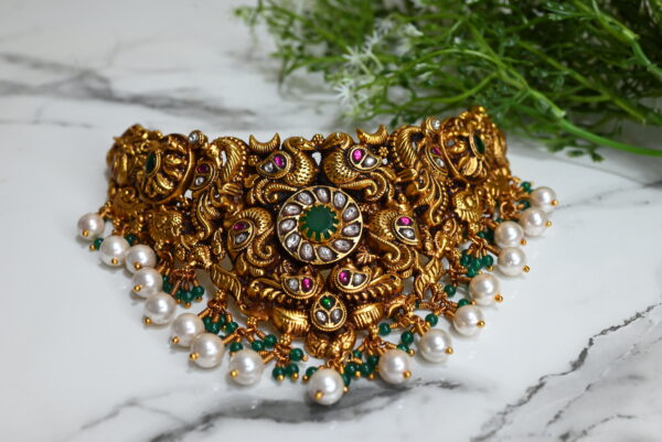 The image showcases an ornate temple-style choker necklace, richly designed in antique gold plating over 925 silver. The centerpiece features a circular green stone surrounded by clear kundan stones. The necklace is adorned with intricate motifs, including peacocks and floral patterns, highlighted with ruby and emerald-colored stones. Hanging below are clusters of white pearls and green beads, adding elegance and movement to the piece. The background is a smooth white marble surface with a sprig of green foliage, enhancing the luxurious and traditional appeal of the jewelry.