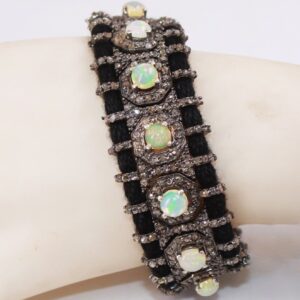 The image shows a detailed close-up of a bracelet. The bracelet features several iridescent opals, each surrounded by numerous small, sparkling diamonds set in what appears to be a dark metal. These gemstone settings are connected and strung together by thick, black thread, creating a textured and contrasting design. The bracelet rests on a smooth, light-colored surface, possibly a display stand or part of a mannequin. 925 Silver Diamond Gemstone Bracelet