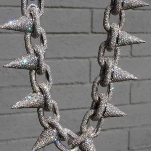 This image shows a bold, high-end hip-hop necklace hanging against a gray brick wall background. The necklace is made of large, thick chain links, each completely encrusted with sparkling diamonds or rhinestones. Attached to every link is a sharp, cone-shaped spike, also fully covered in shimmering stones. The entire piece glitters intensely under the light, giving off rainbow reflections and a highly luxurious, eye-catching appearance. The design blends rugged toughness with flashy extravagance, typical of statement jewelry in hip-hop fashion. "925 Silver Hip-Hop Necklace"