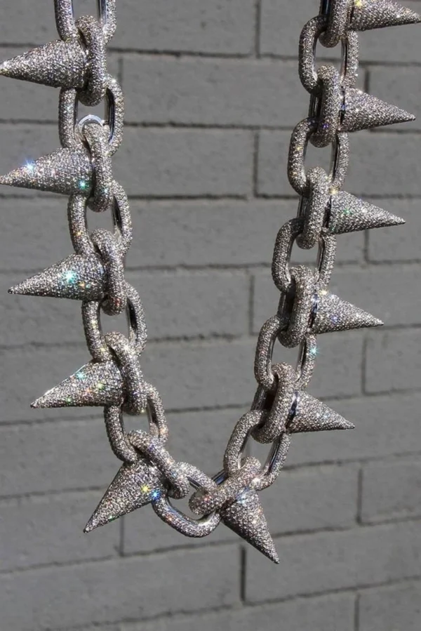This image shows a bold, high-end hip-hop necklace hanging against a gray brick wall background. The necklace is made of large, thick chain links, each completely encrusted with sparkling diamonds or rhinestones. Attached to every link is a sharp, cone-shaped spike, also fully covered in shimmering stones. The entire piece glitters intensely under the light, giving off rainbow reflections and a highly luxurious, eye-catching appearance. The design blends rugged toughness with flashy extravagance, typical of statement jewelry in hip-hop fashion. "925 Silver Hip-Hop Necklace"