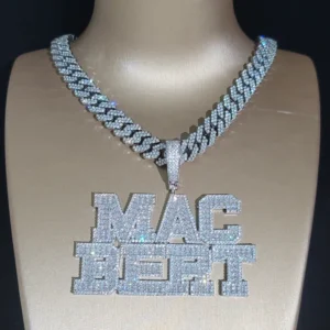 The image shows a luxurious men's necklace displayed on a mannequin. The necklace features a thick Cuban link chain, completely encrusted with sparkling stones, likely cubic zirconia or diamonds. Hanging from the chain is a large, bold pendant that spells out "MAC PORT" in block letters, also fully covered in shimmering stones. The jewelry has a brilliant, high-end shine, typical of hip-hop style, emphasizing wealth, confidence, and bold fashion. The background is dark, helping the necklace's sparkle stand out even more vividly.